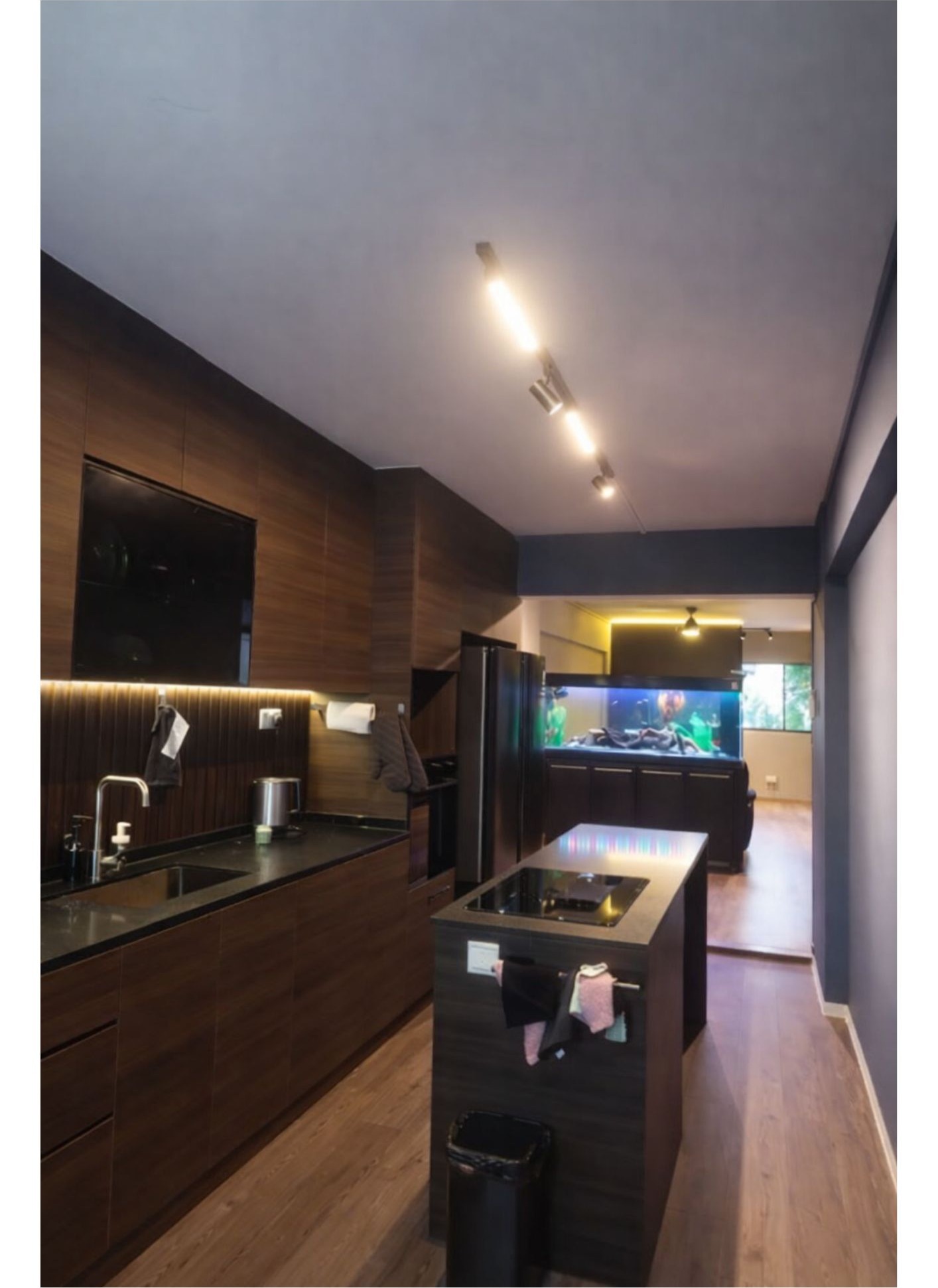 Modern luxe 5-room HDB kitchen with island counter and dark timber cabinetry, viewed from corridor with fish tank in living area beyond